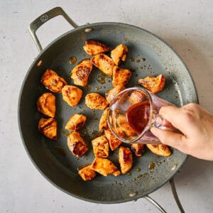 pouring thick korean bbq sauce over cooked cubed chicken pieces in a pan.