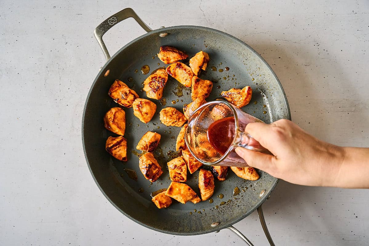 pouring thick korean bbq sauce over cooked cubed chicken pieces in a pan.