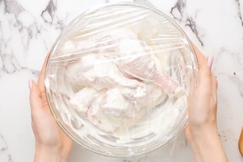 Covering a bowl of chicken with plastic wrap to marinate.