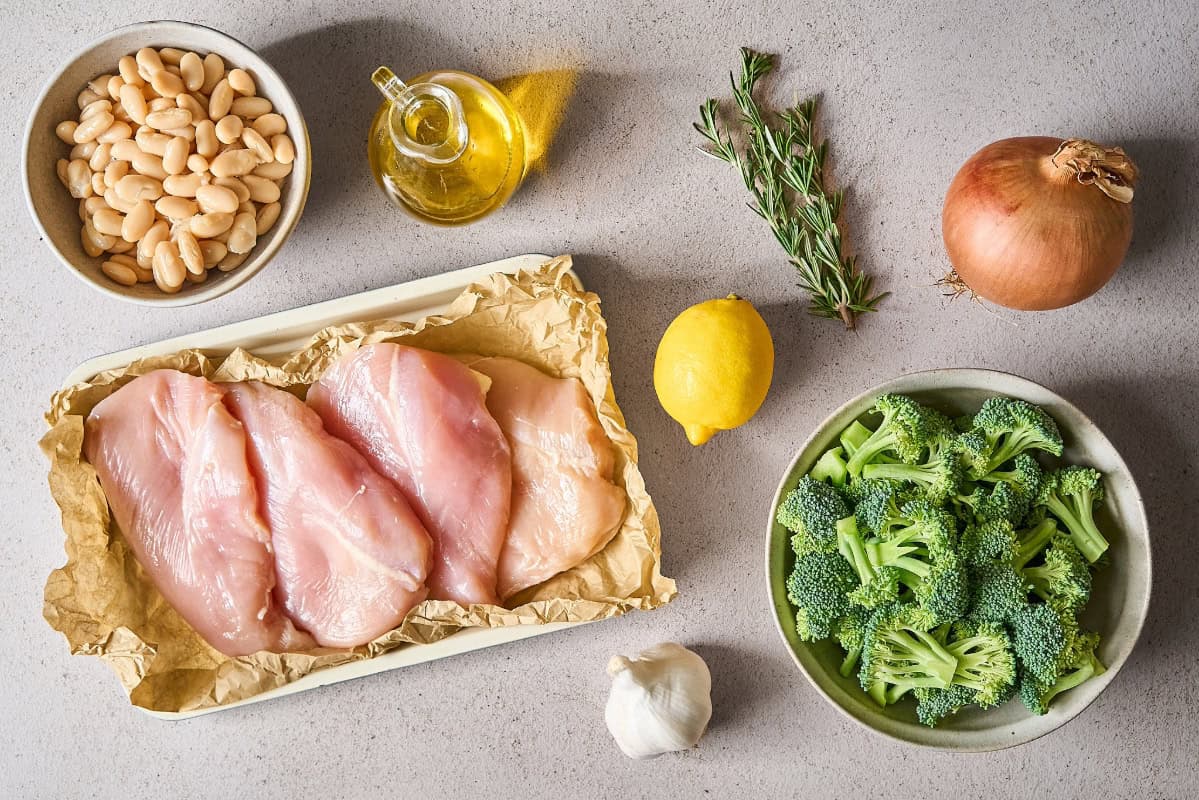 ingredients for lemon chicken skillet meal: chicken breasts, white beans, rosemary, broccoli, onion, lemon, and garlic.