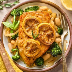 overhead view of a chicken breast topped with lemon slices on a bed of white beans and broccoli with a fork.