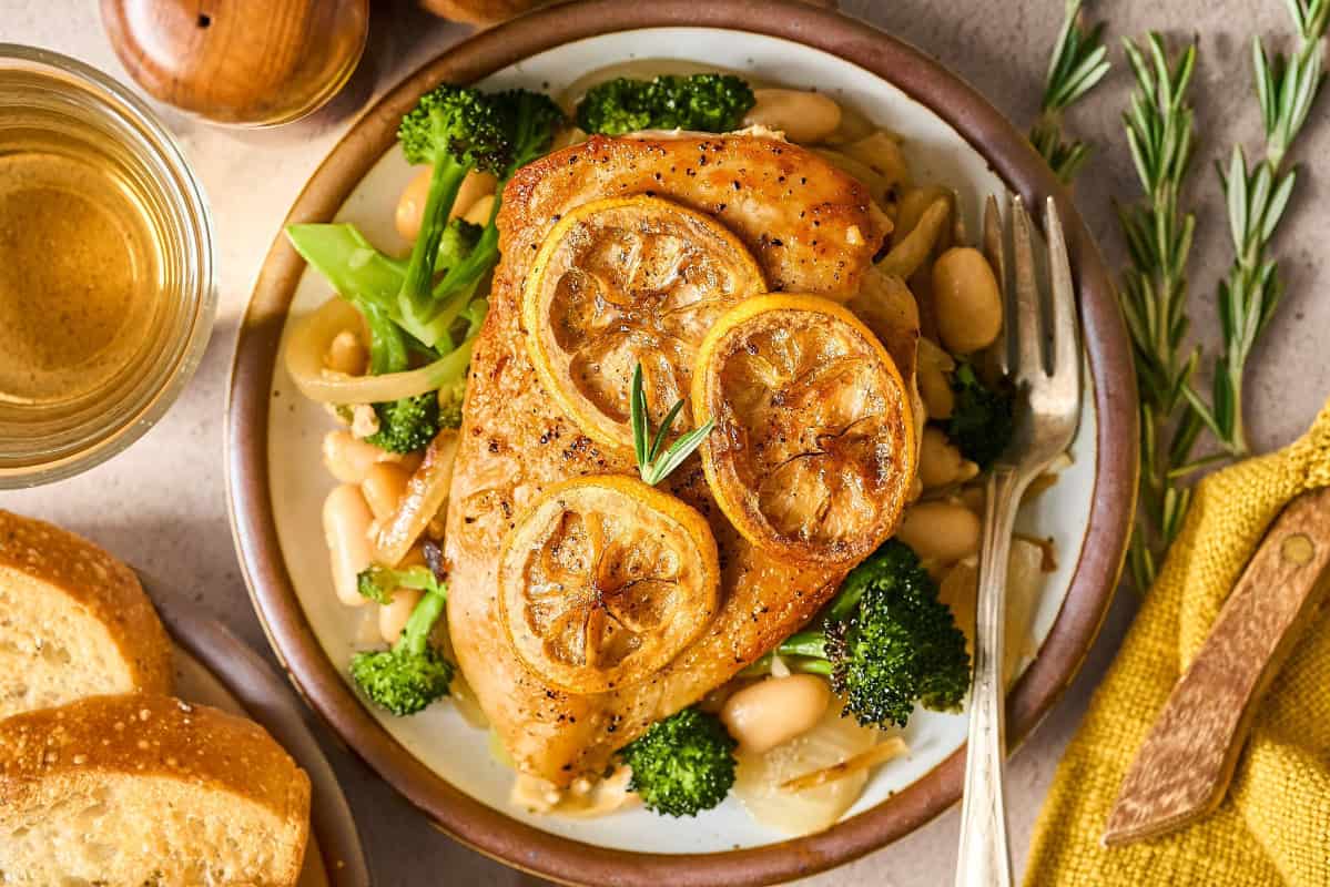 overhead view of a rosemary chicken breast topped with lemon slices on a bed of white beans and broccoli with a fork.