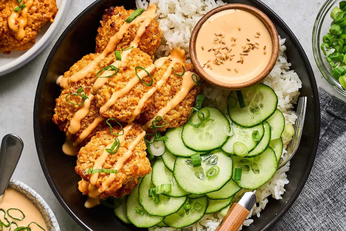 overhead view of a serving of baked bang bang chicken in a black bowl with rice, cucumbers, and bang bang sauce.