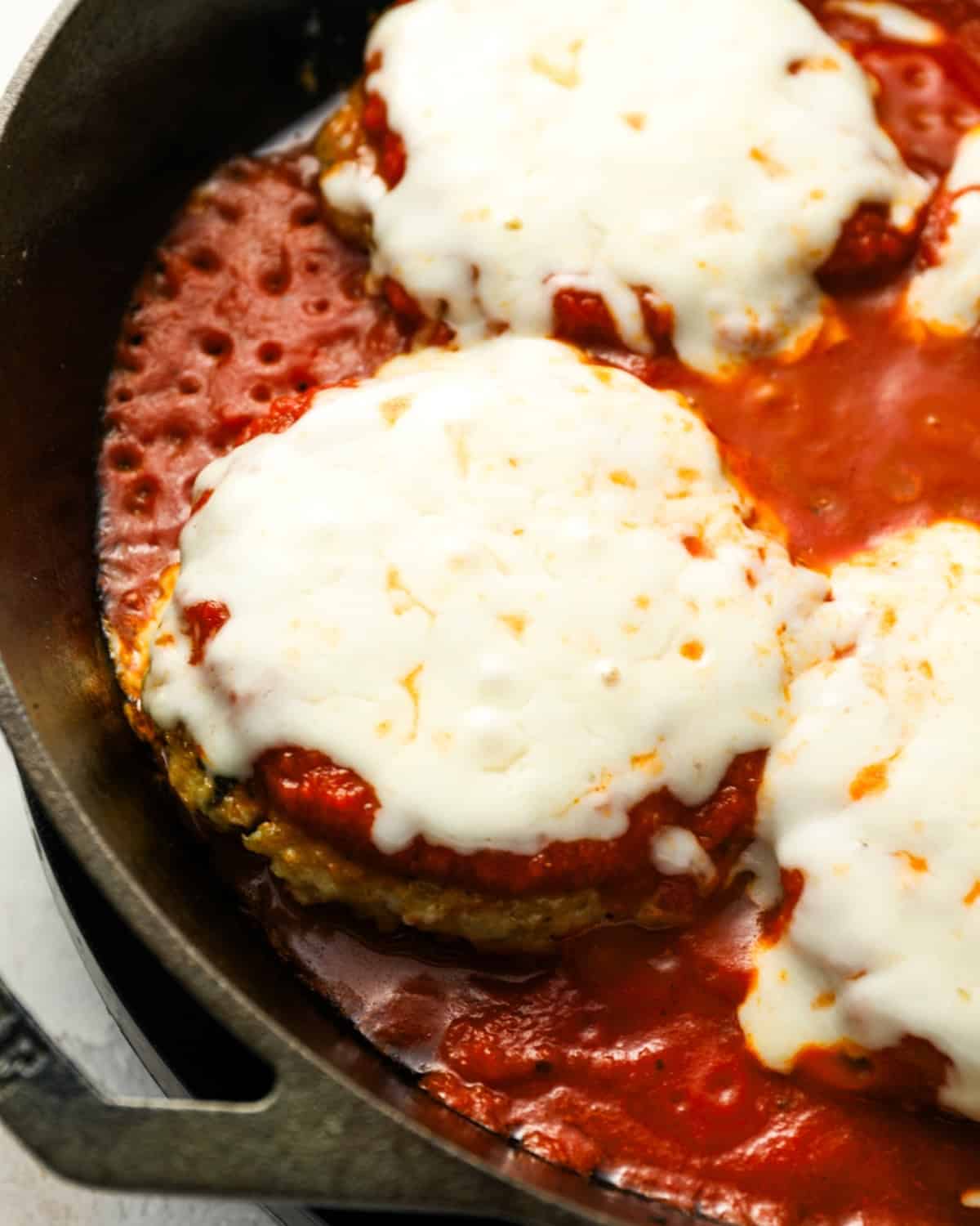 Breaded chicken burger patties, coated in marinara sauce and cheese, in a cast iron skillet.