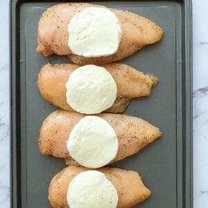 Chicken breasts topped with mozzarella, lined up on a sheet pan.
