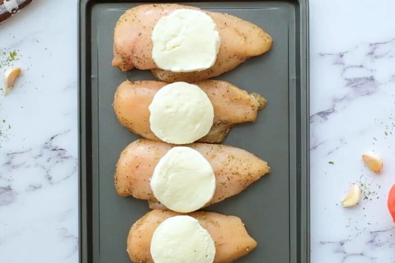 Chicken breasts topped with mozzarella, lined up on a sheet pan.