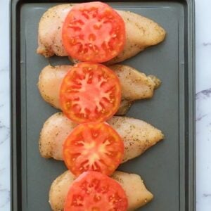 Chicken breasts topped with tomatoes, lined up on a sheet pan.