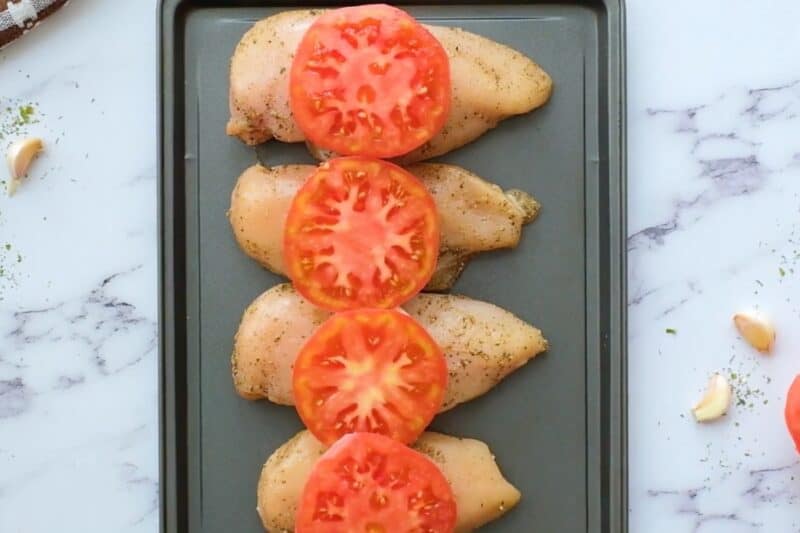 Chicken breasts topped with tomatoes, lined up on a sheet pan.