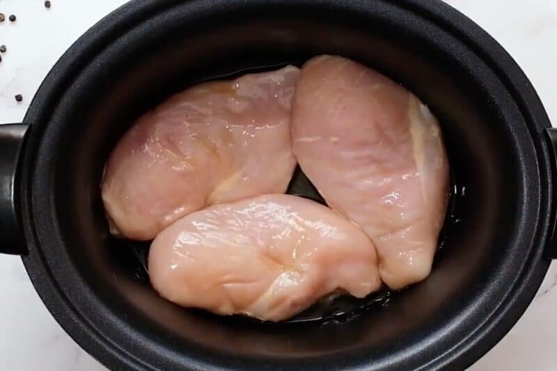 Raw chicken breasts coated in olive oil, in a slow cooker.