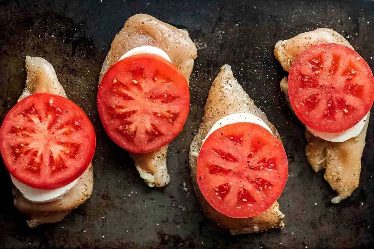 Chicken breasts on a sheet pan, topped with tomatoes and mozzarella, before baking.