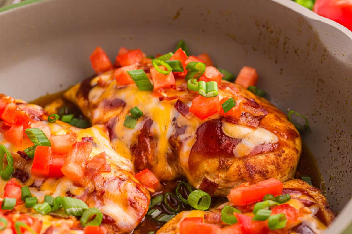 Cheesy BBQ chicken breasts in a skillet, garnished with tomatoes, green onions, and bacon.