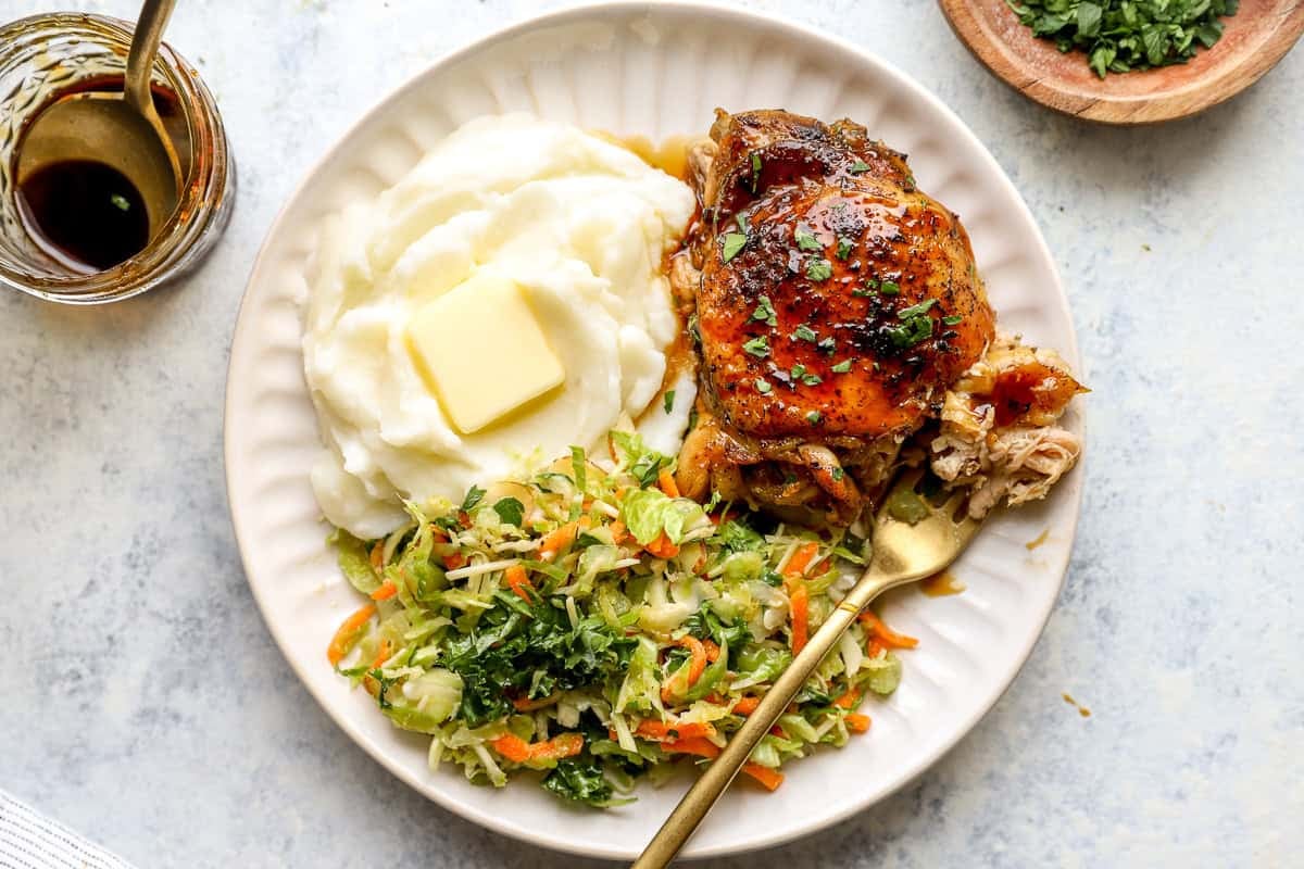 A serving of Crockpot chicken thighs, on a plate of mashed potatoes and slaw.