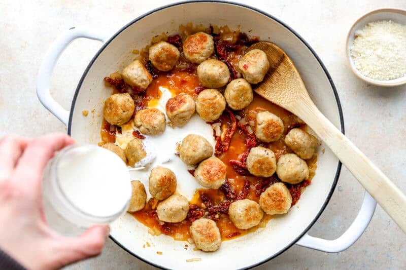 Cream being poured into a skillet with meatballs.
