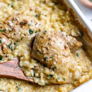 Wooden spoon lifting a chicken thigh out of a dish of creamy rice casserole.