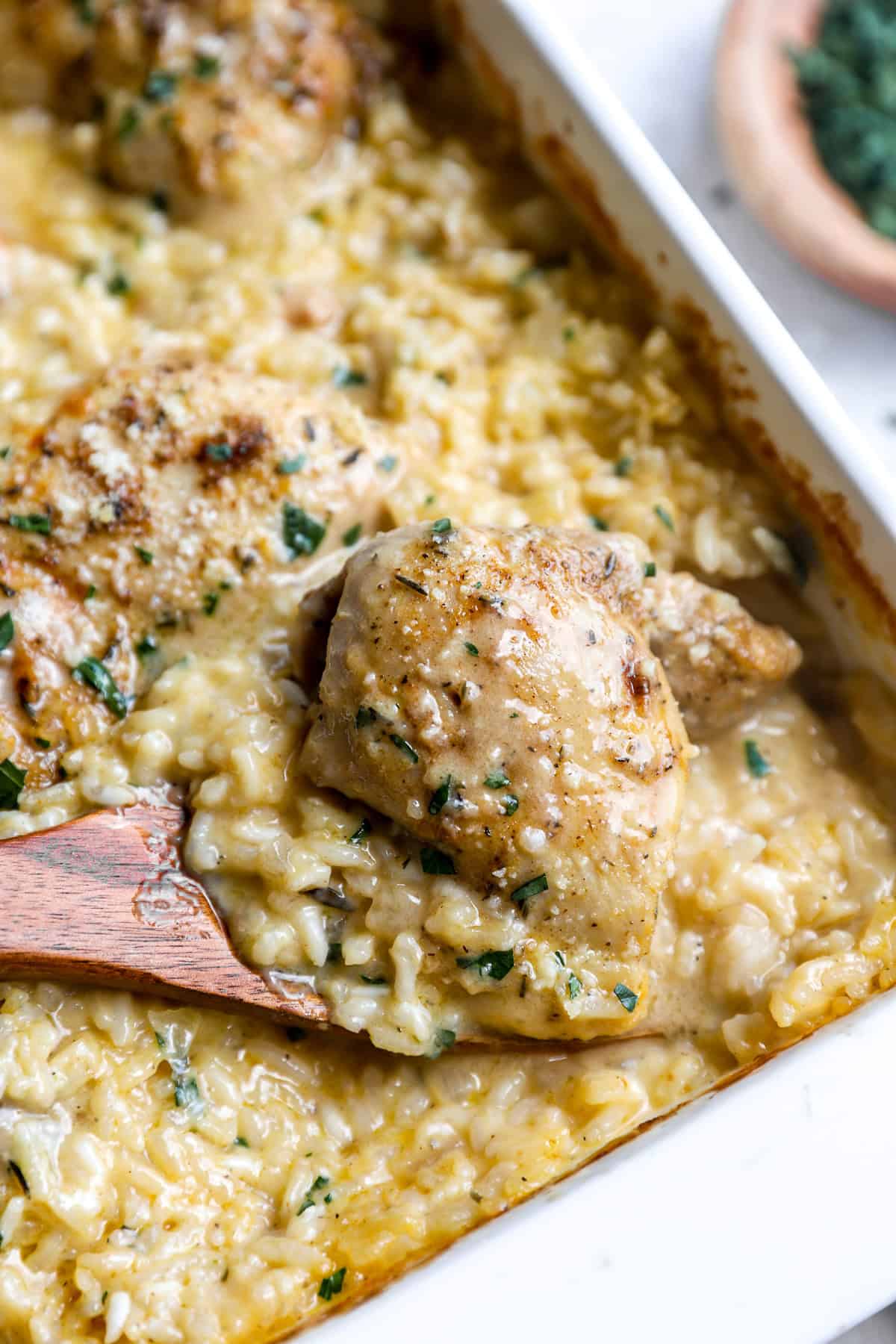 Wooden spoon lifting a chicken thigh out of a dish of creamy rice casserole.