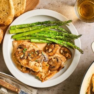Chicken Diane on a white plate with a side of asparagus and a knife surrounded by crusty bread and a cup of brandy.
