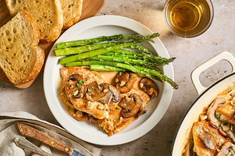 Chicken Diane on a white plate with a side of asparagus and a knife surrounded by crusty bread and a cup of brandy.