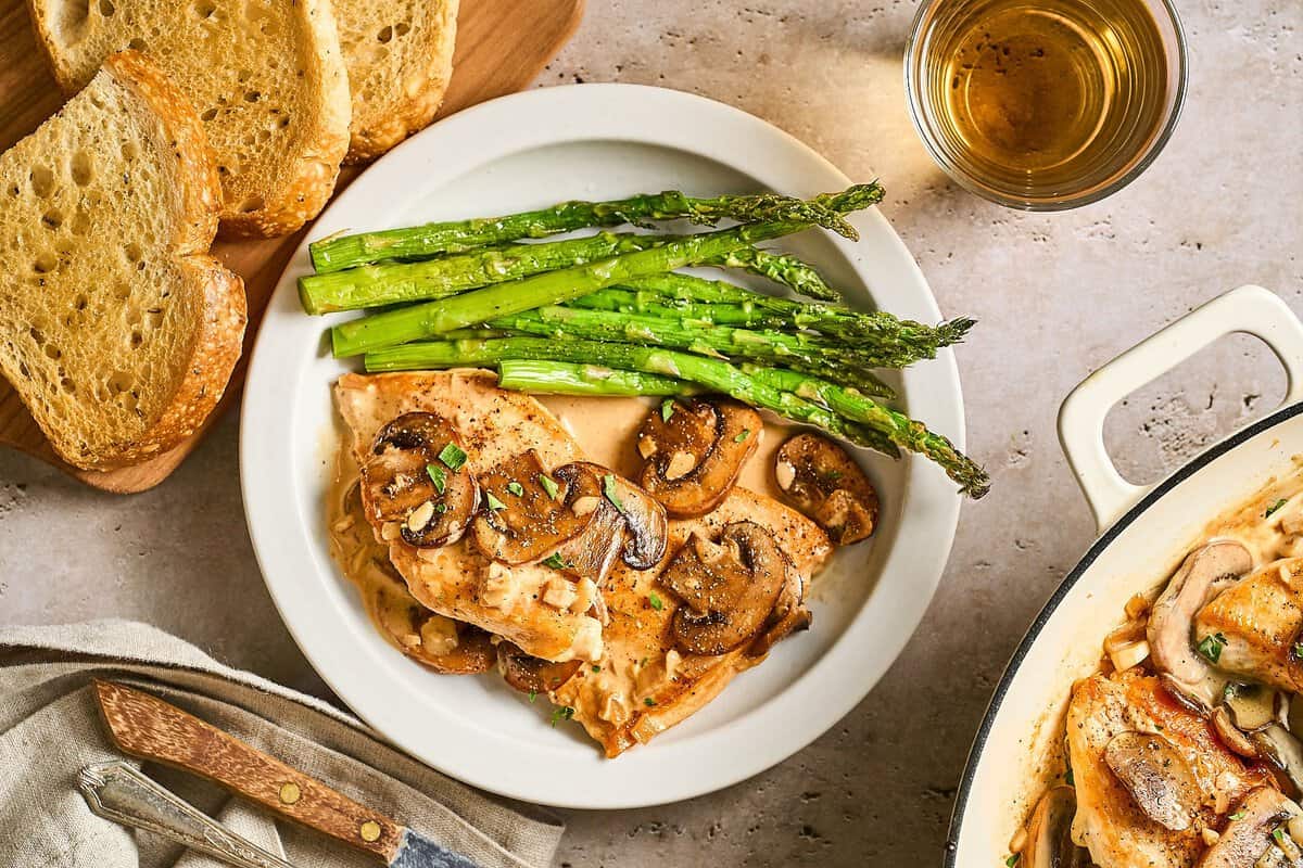 Chicken Diane on a white plate with a side of asparagus and a knife surrounded by crusty bread and a cup of brandy.