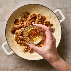 A hand holding a small bowl of brandy over a skillet of browned mushrooms.