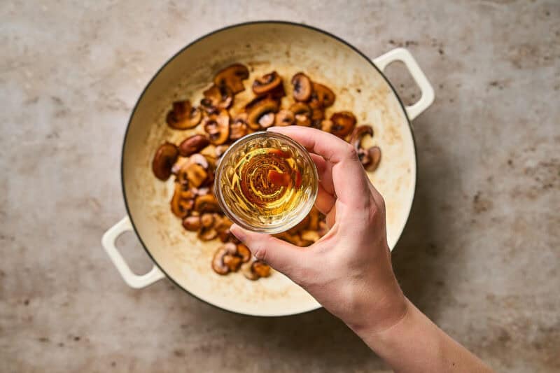 A hand holding a small bowl of brandy over a skillet of browned mushrooms.