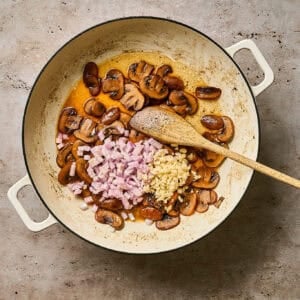 Stirring minced shallot and garlic into a skillet of browned mushrooms with a wooden spoon.