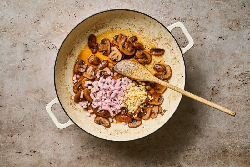 Stirring minced shallot and garlic into a skillet of browned mushrooms with a wooden spoon.