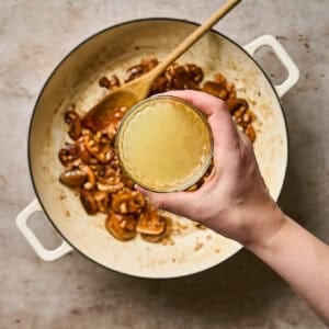 A hand holding a small bowl of chicken broth over a skillet of browned mushrooms.