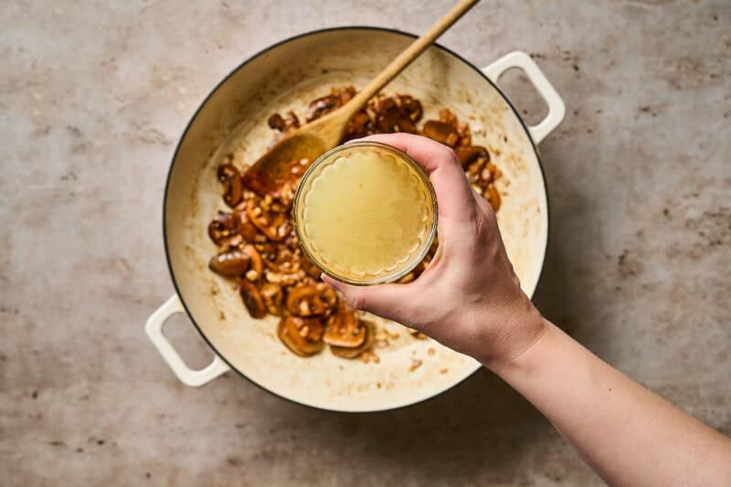 A hand holding a small bowl of chicken broth over a skillet of browned mushrooms.