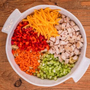 Diced chicken, shredded cheddar cheese, diced celery, red bell pepper, and chopped carrots in a large white mixing bowl.
