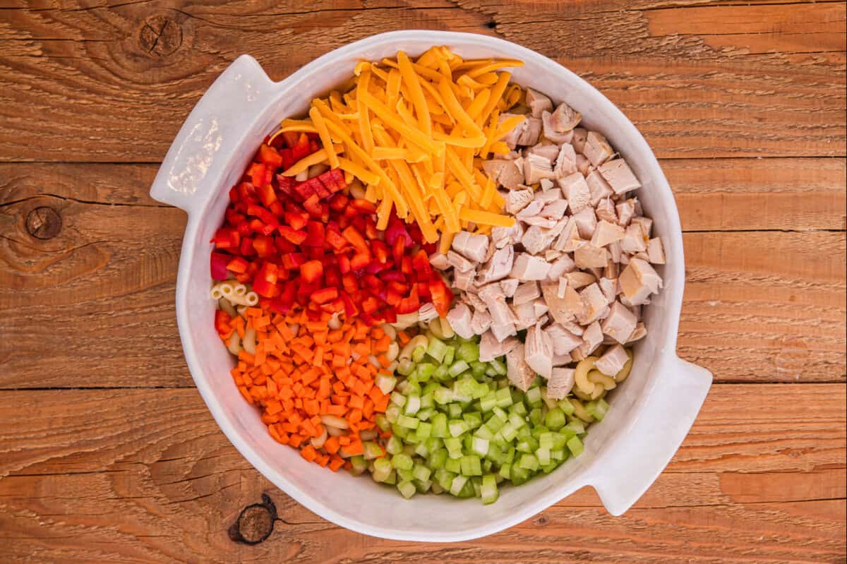 Diced chicken, shredded cheddar cheese, diced celery, red bell pepper, and chopped carrots in a large white mixing bowl.