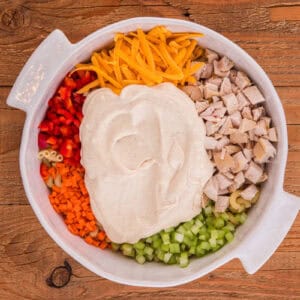 Creamy dressing added to a bowl filled with macaroni, chicken, cheese, and veggies.