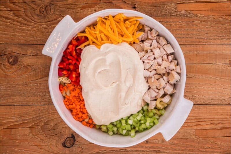 Creamy dressing added to a bowl filled with macaroni, chicken, cheese, and veggies.