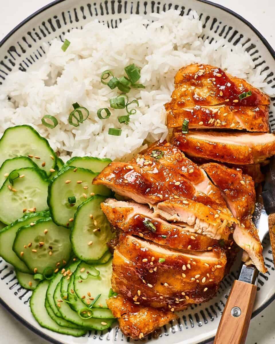 A plate of shoyu glazed chicken thighs with white rice and cucumbers.