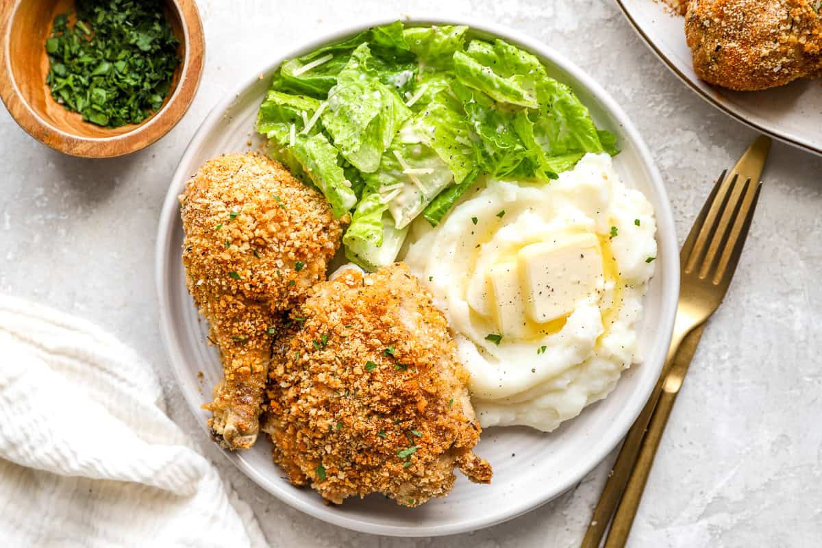 A serving of copycat Shake and Bake chicken legs on a plate of mashed potatoes and salad.