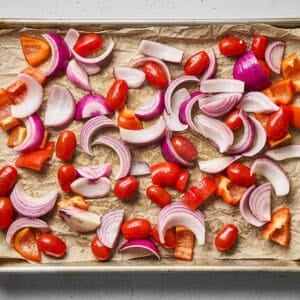 Red onion slices, chopped red bell pepper, and grape tomatoes arranged on a sheet pan.