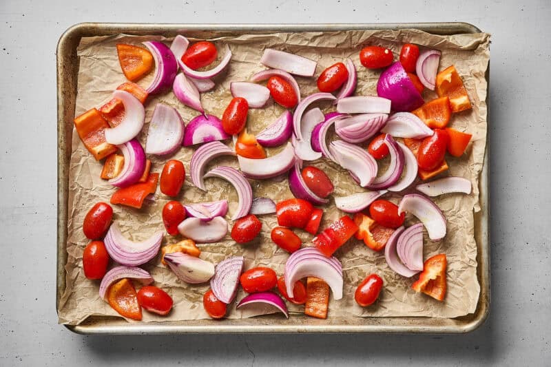 Red onion slices, chopped red bell pepper, and grape tomatoes arranged on a sheet pan.