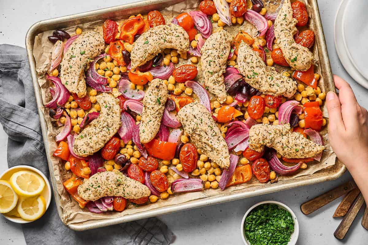 Hand grabbing a sheet pan filled with chicken tenders, chickpeas, tomatoes, olives, and onion.