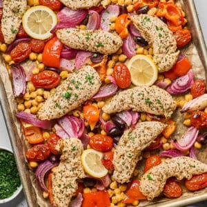 A sheet pan filled with Greek marinated chicken tenders, chickpeas, olives, tomatoes, and red onion.
