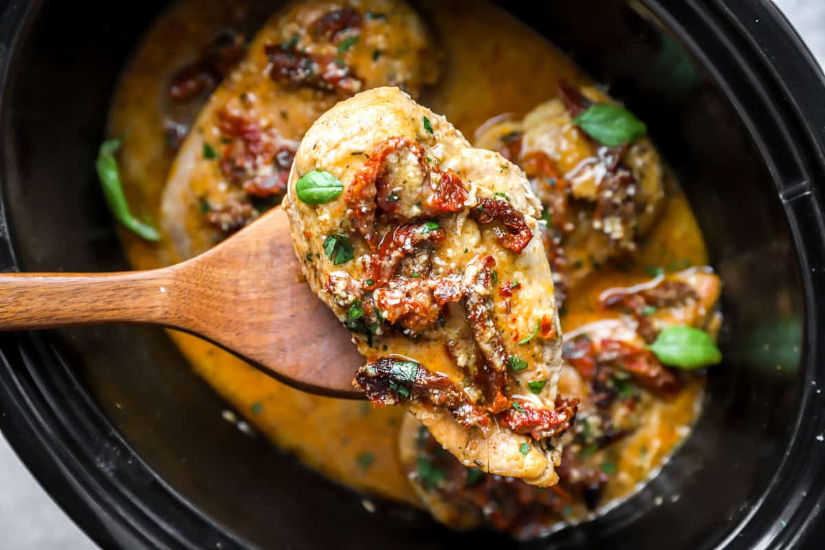 Wooden spoon holding up a chicken breast topped with sun-dried tomatoes.